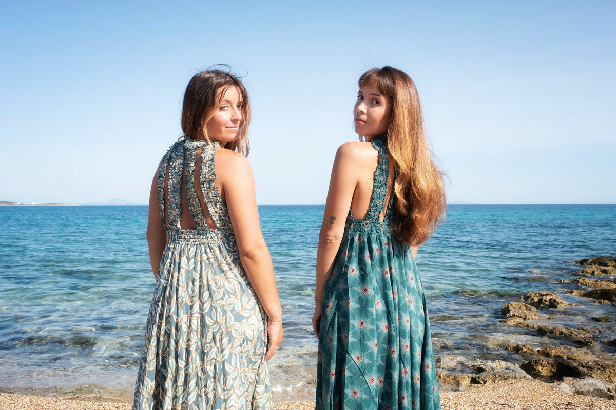 Two women in printed maxi dresses on a pebble beach, looking back over their shoulders at the sea