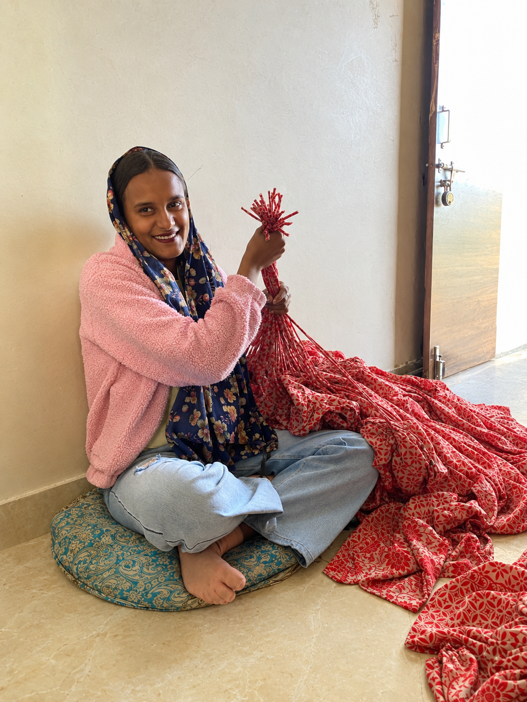 A smiling woman sitting on a floor cushion, holding a bundle of red printed drawstrings