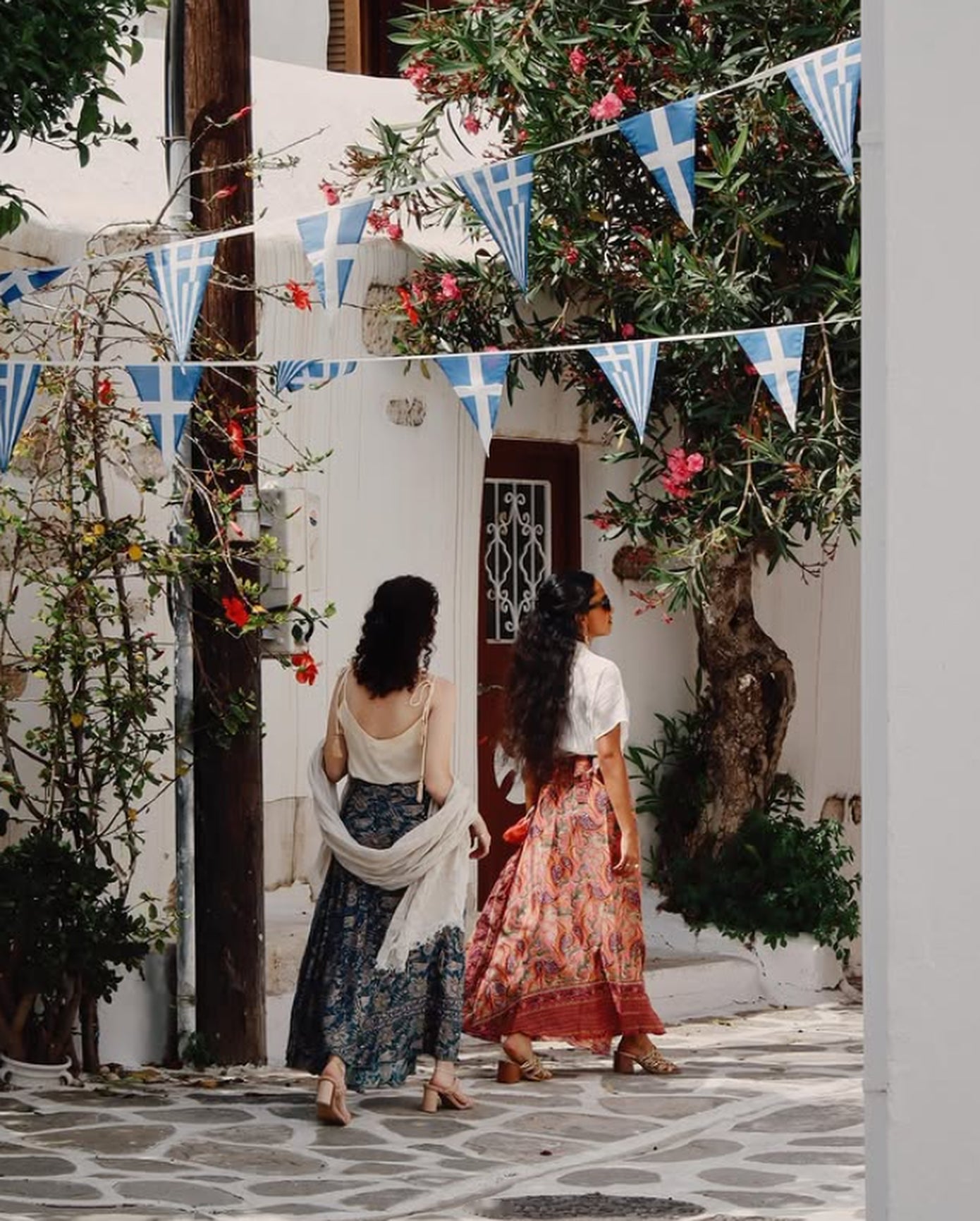 Two women walking away down a Paros lane under Greek flags and oleander